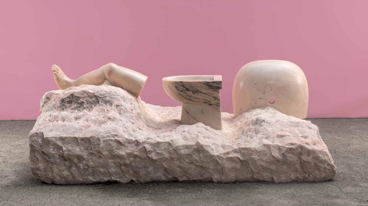 Sculpture on marble slab with realistic human leg and toilet, set against a pink background, showcasing artistic juxtaposition.