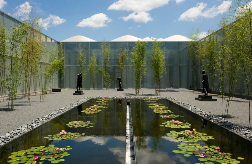 Modern outdoor sculpture garden with reflecting pool, surrounded by bamboo and statues under a cloudy sky.