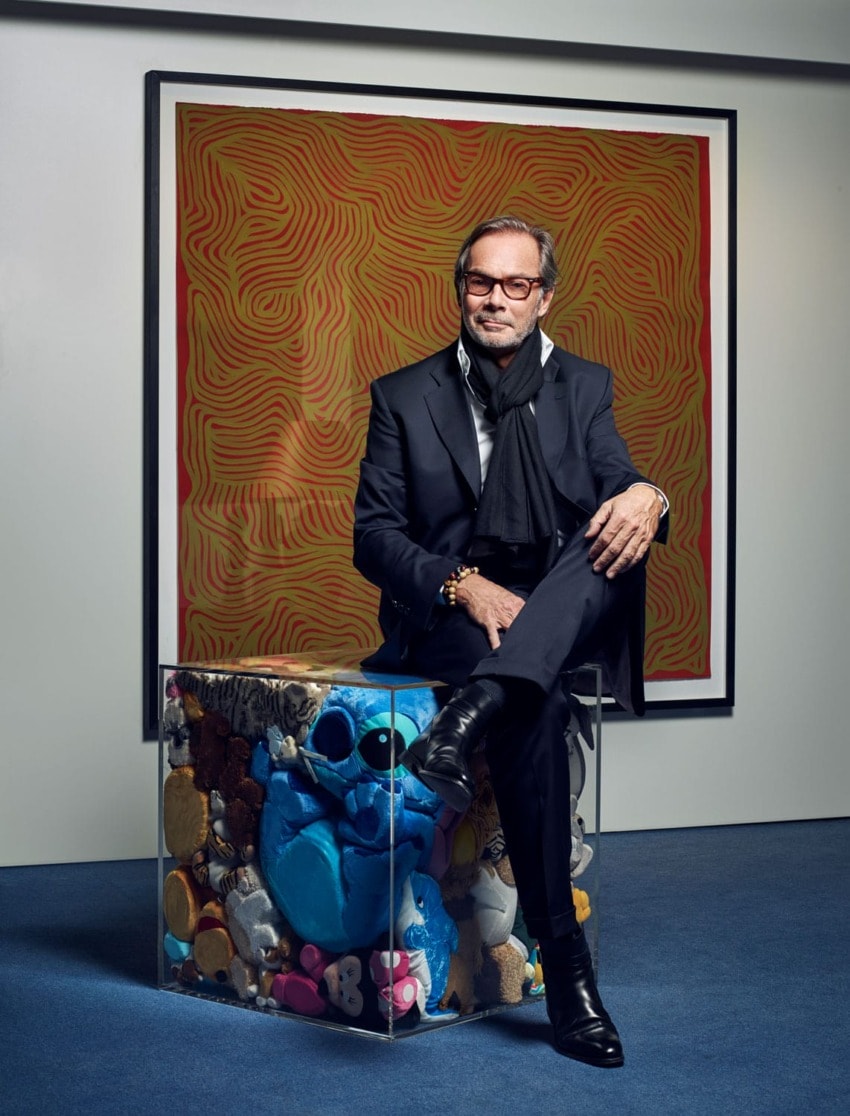 Person sitting on a transparent cube with objects inside, in front of abstract red and black artwork, wearing a black outfit.