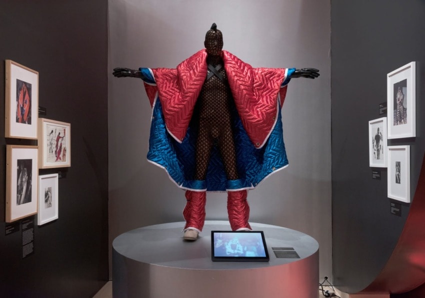 Mannequin in colorful quilted costume on display among framed artworks in an exhibit.