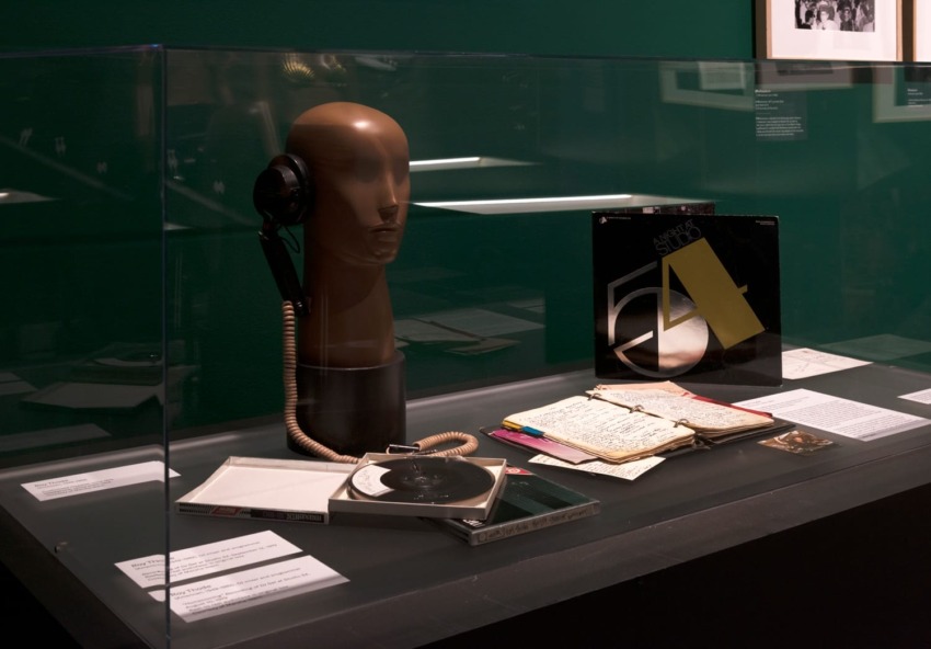 Museum exhibit with mannequin head wearing headphones, vinyl record, lined paper with notes, and colorful pens on display.