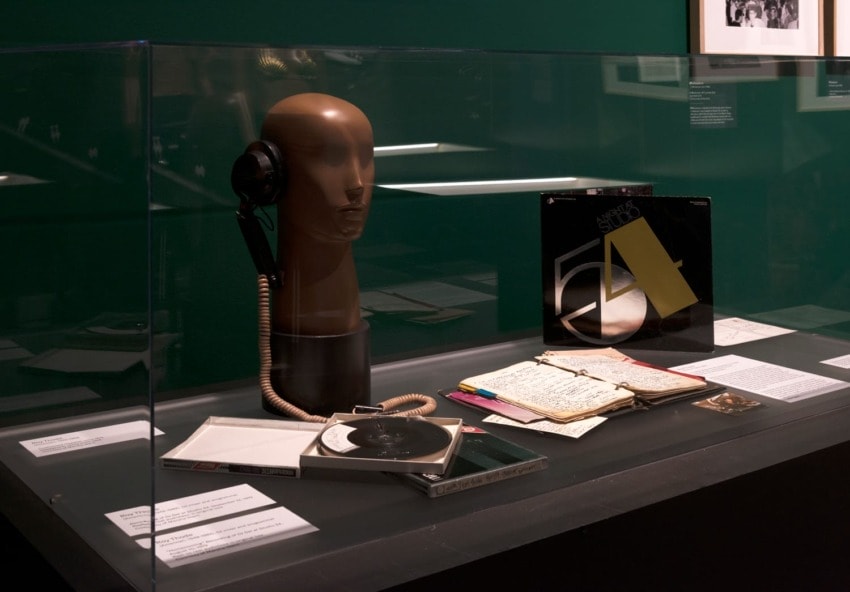 Museum exhibit with mannequin head wearing headphones, vinyl record, lined paper with notes, and colorful pens on display.