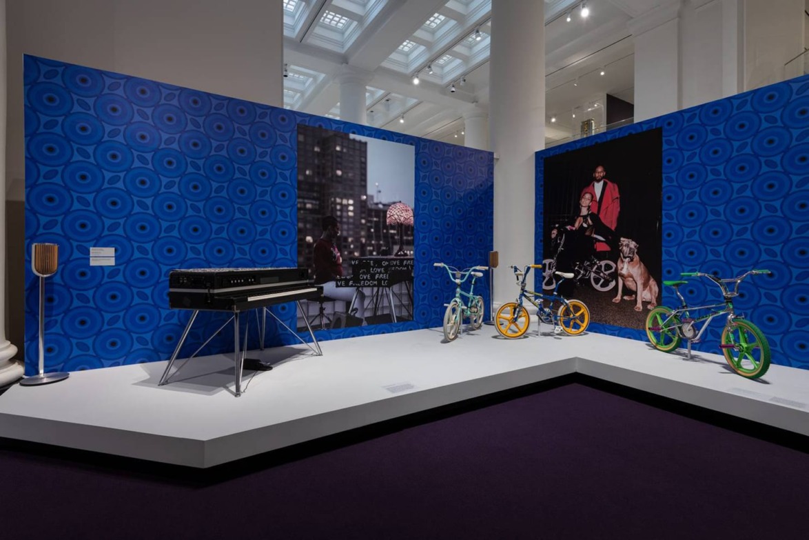 Exhibit showcasing a keyboard, colorful bicycles, and large cityscape portraits on blue patterned walls in a gallery setting.