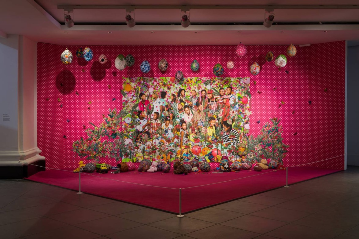 Colorful art exhibit with a vibrant collage on a pink wall, featuring people, patterns, and hanging ornaments.