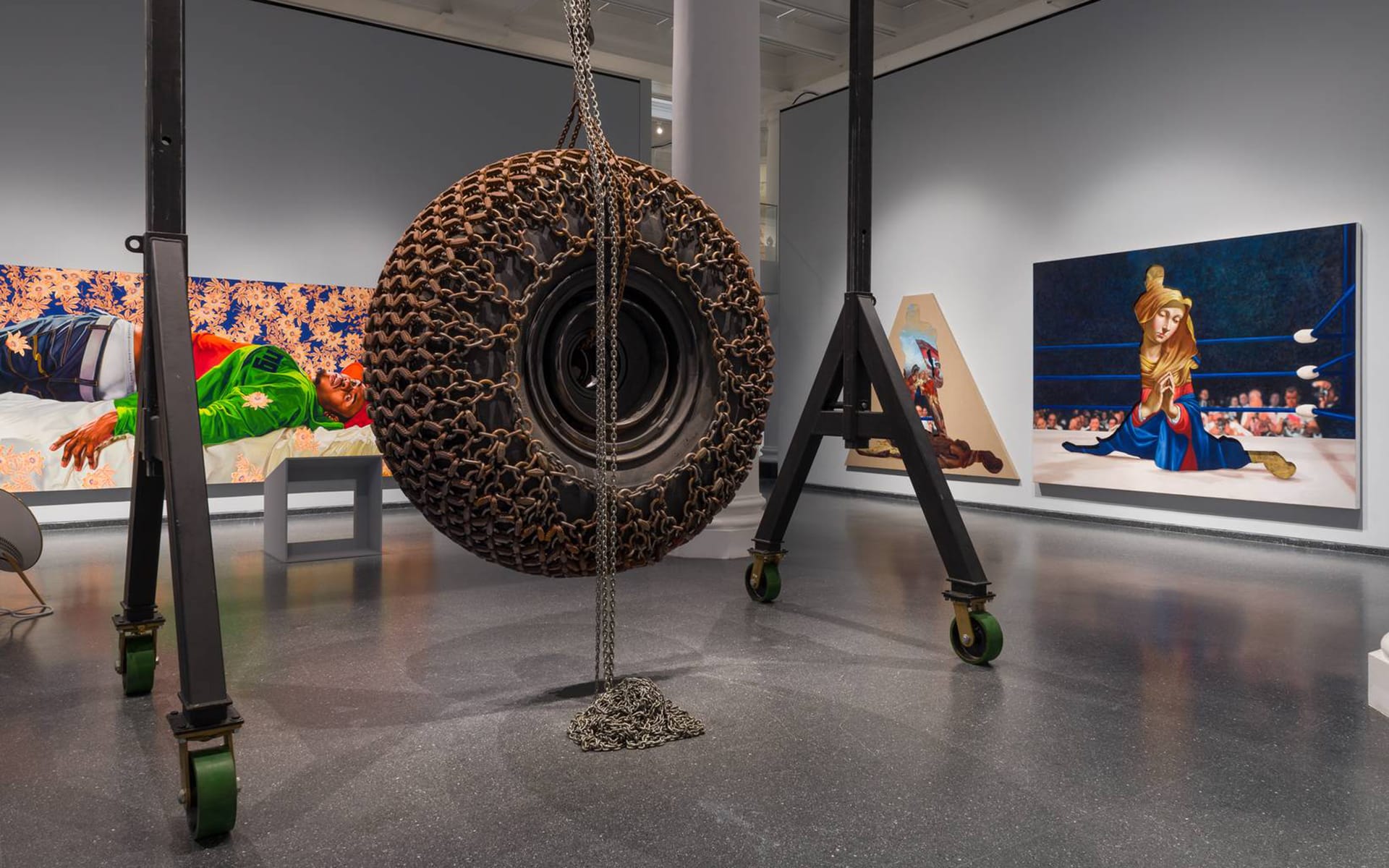 Art exhibit with a large sculptural tire and colorful paintings on walls, featuring various scenes and figures in a gallery setting.