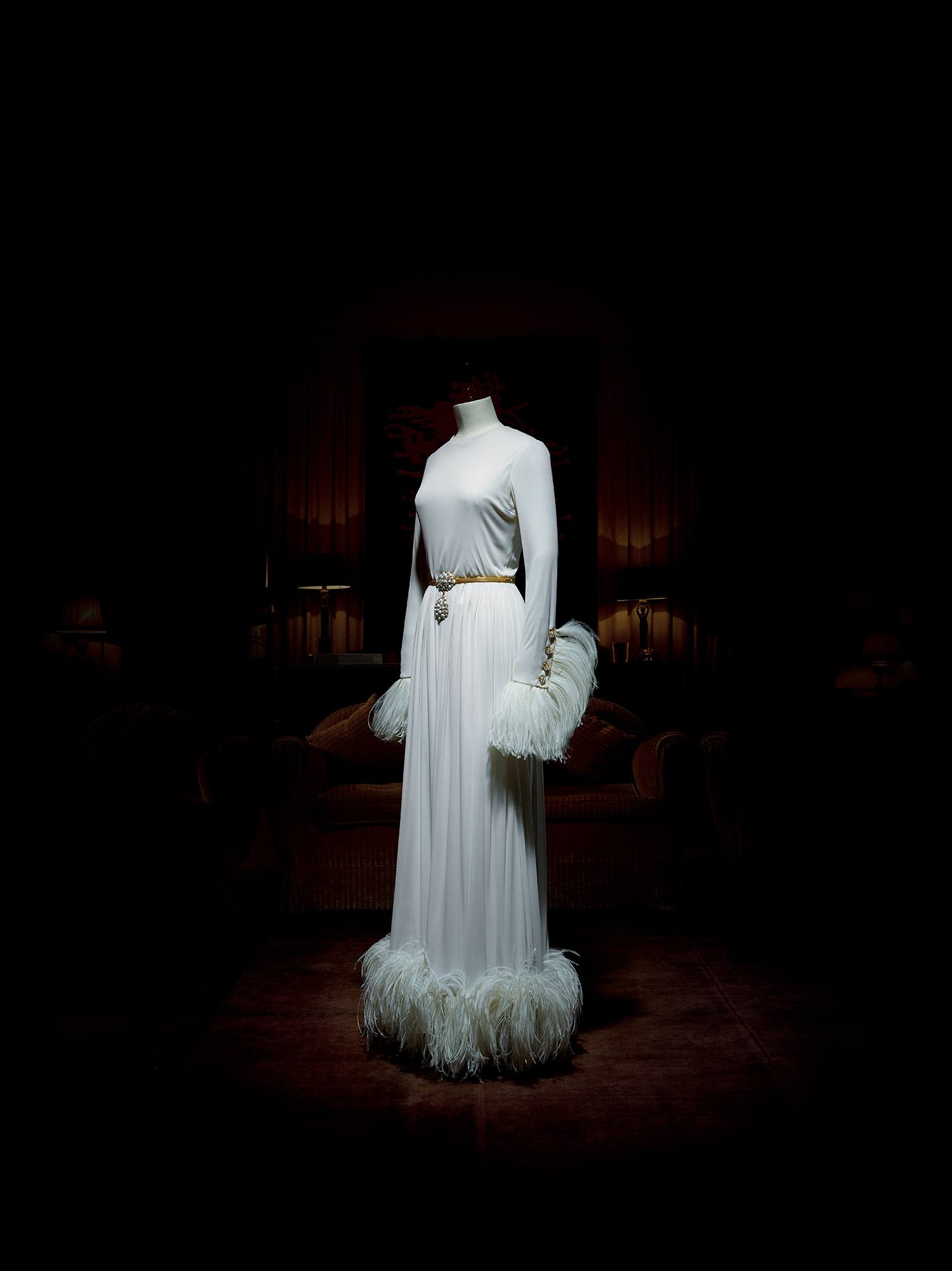 White floor-length gown with feathered cuffs and hem on a mannequin in a dimly lit room.