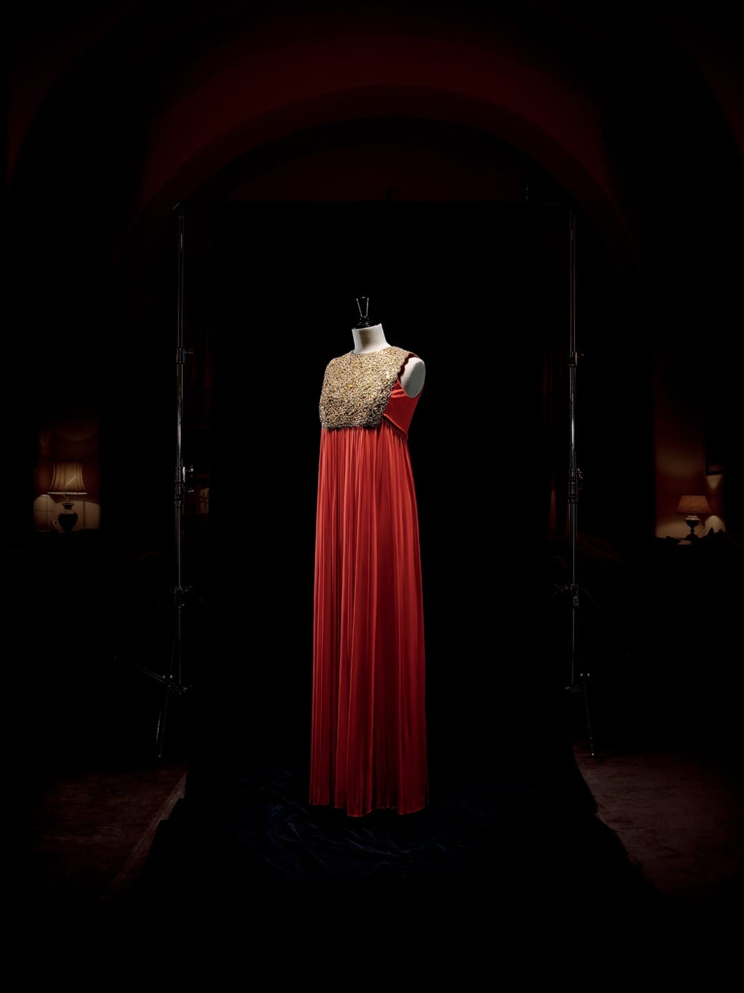 Red sleeveless evening gown with a gold embellished bodice displayed on a mannequin in a dimly lit room.