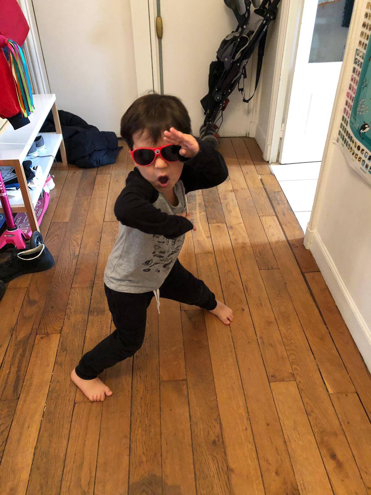 Child wearing red sunglasses striking a playful pose on a wooden floor indoors.