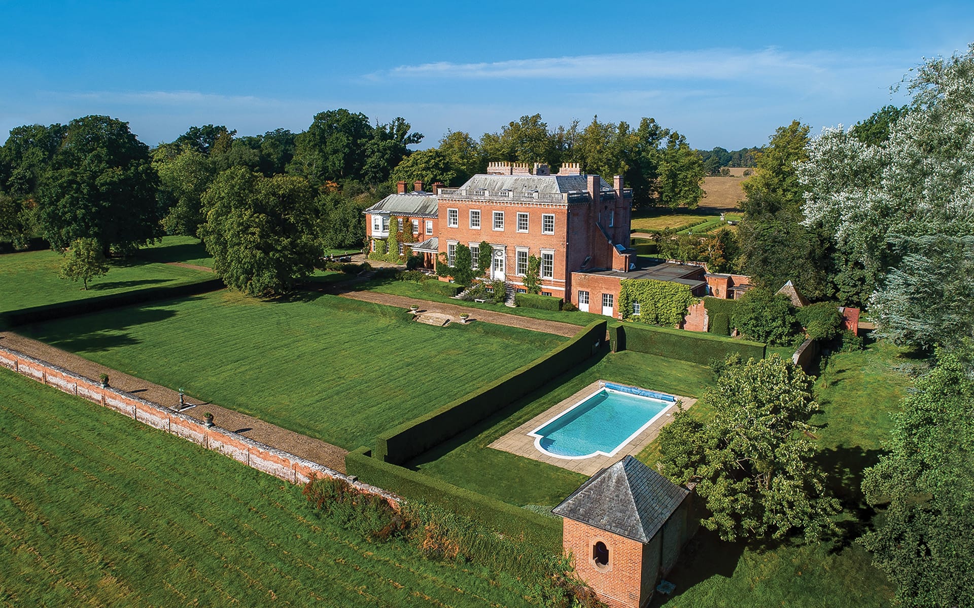 Aerial view of a large brick mansion with a swimming pool and expansive green lawns surrounded by trees.