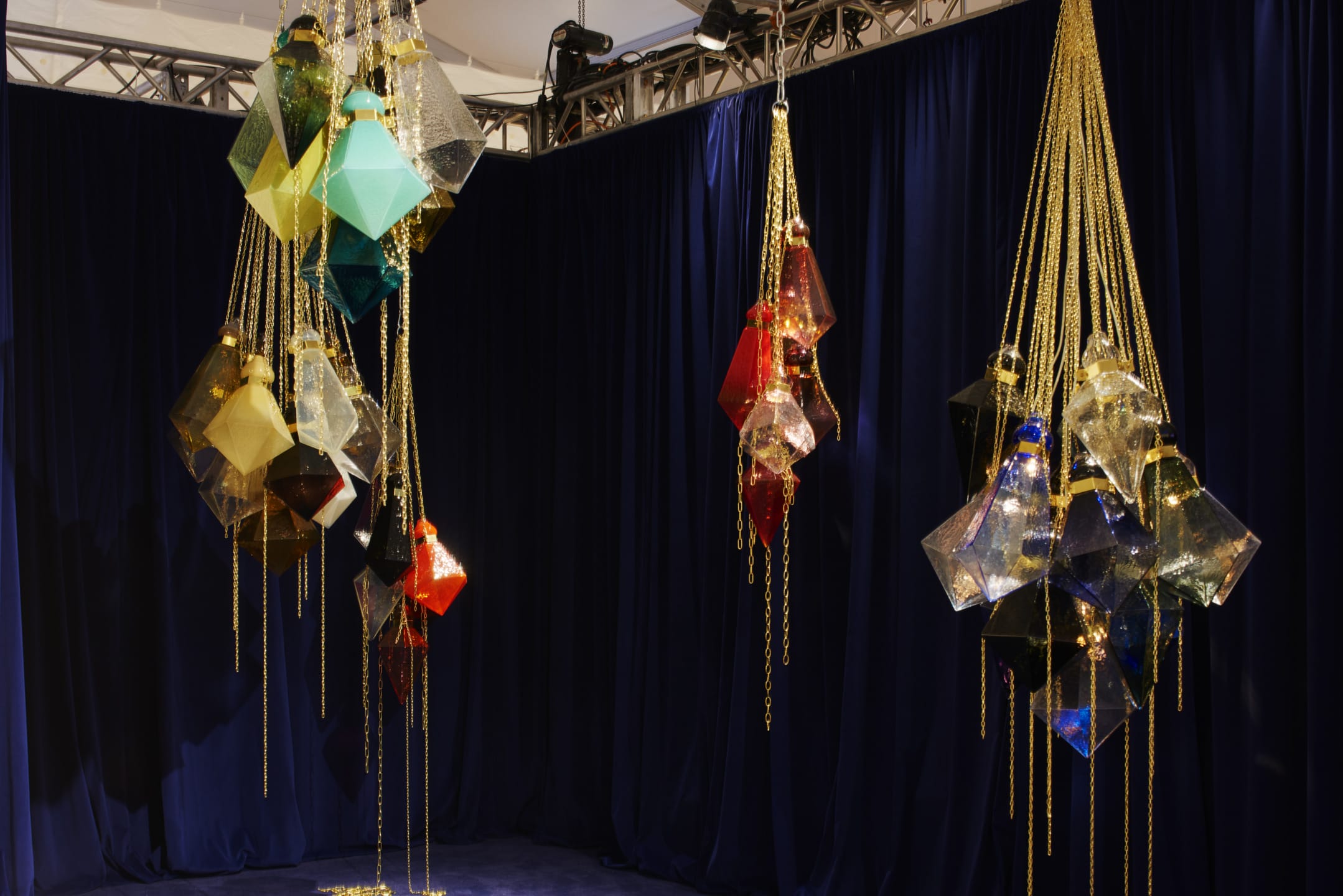 Colorful geometric chandeliers with hanging gold chains against a dark blue curtain backdrop.