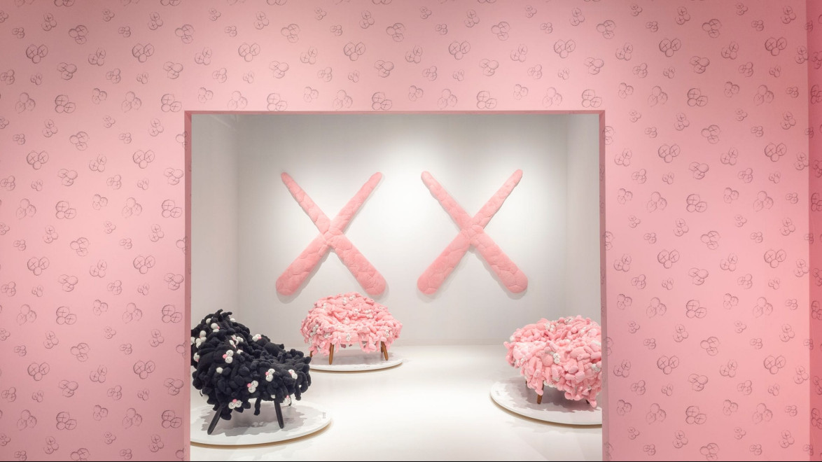 Gallery exhibit with large pink crossed shapes on wall and clusters of pink and black sculptures on display stands.