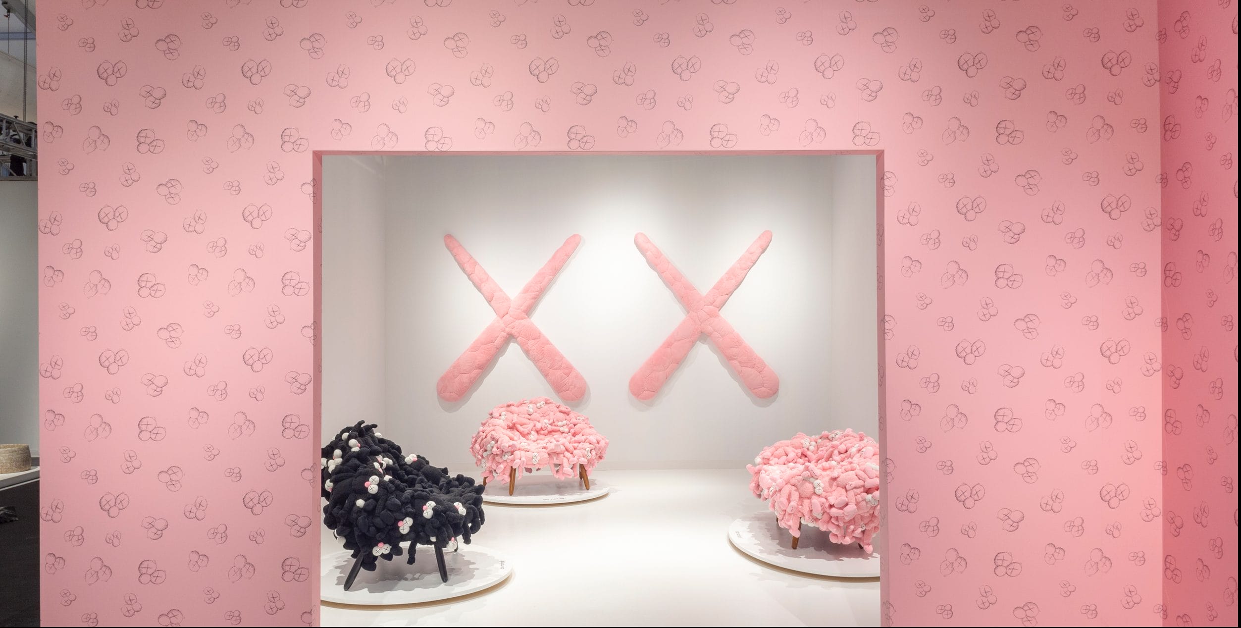 Gallery exhibit with large pink crossed shapes on wall and clusters of pink and black sculptures on display stands.