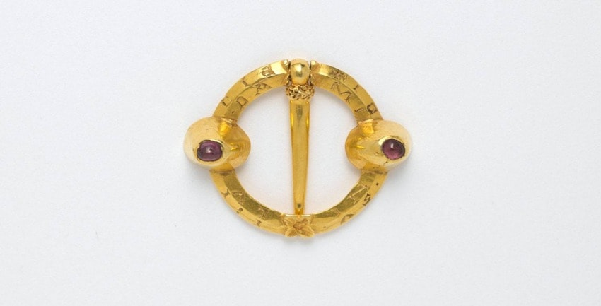 Medieval-style gold brooch with circular design, featuring two red gems on either side of a central pin.