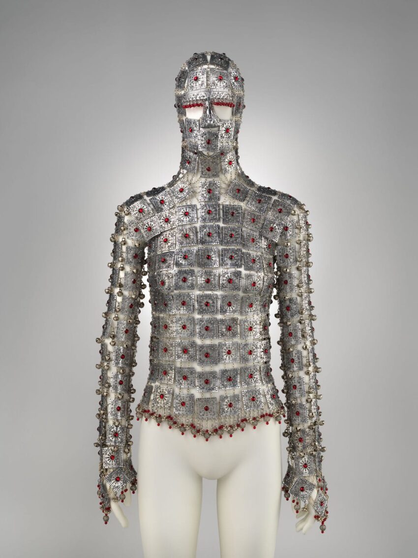 Chainmail-style suit with intricate geometric patterns and red accents displayed on a mannequin against a gray background.