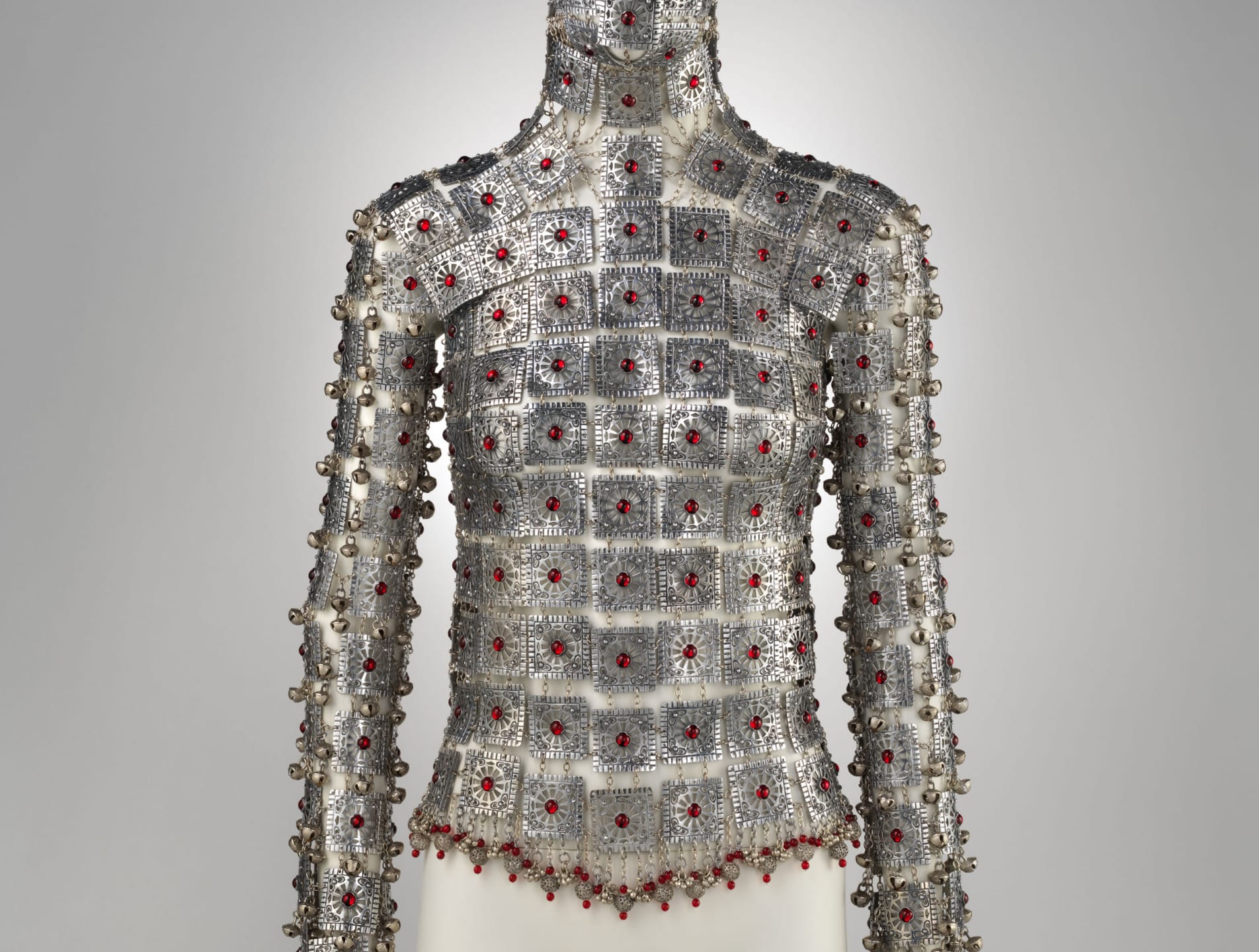 Intricate metallic armor-like top adorned with red gemstones displayed on a mannequin against a neutral background.
