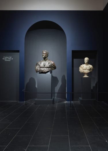 Two sculpted busts displayed in a museum with dark blue walls and a spotlight highlighting their artistic details.