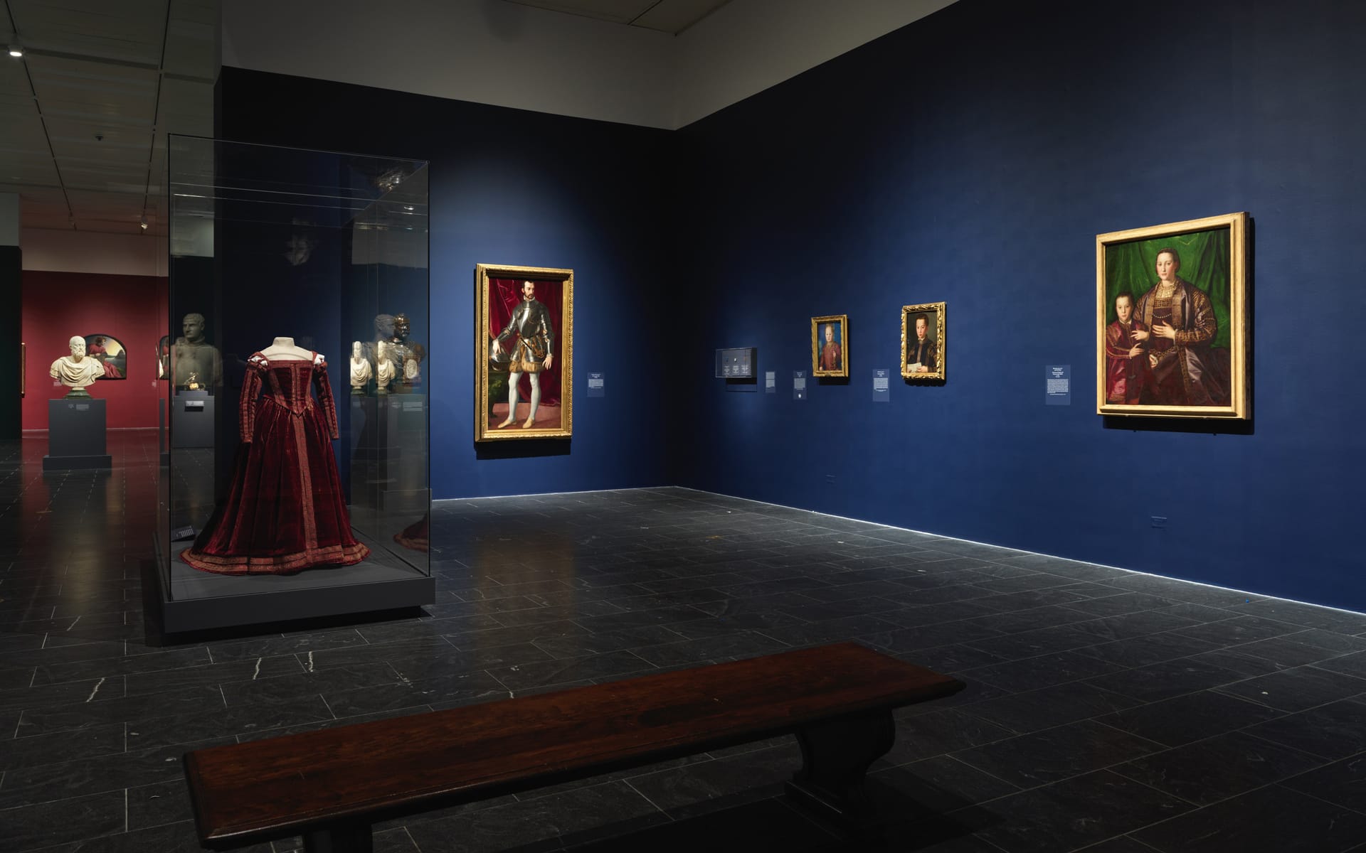 Art gallery with classical portraits on dark walls, showcasing paintings, sculptures, and a red dress display.