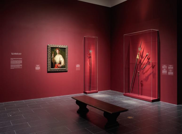 Art gallery with a portrait of a man, display cases of swords on a red wall, and a wooden bench on a stone floor.