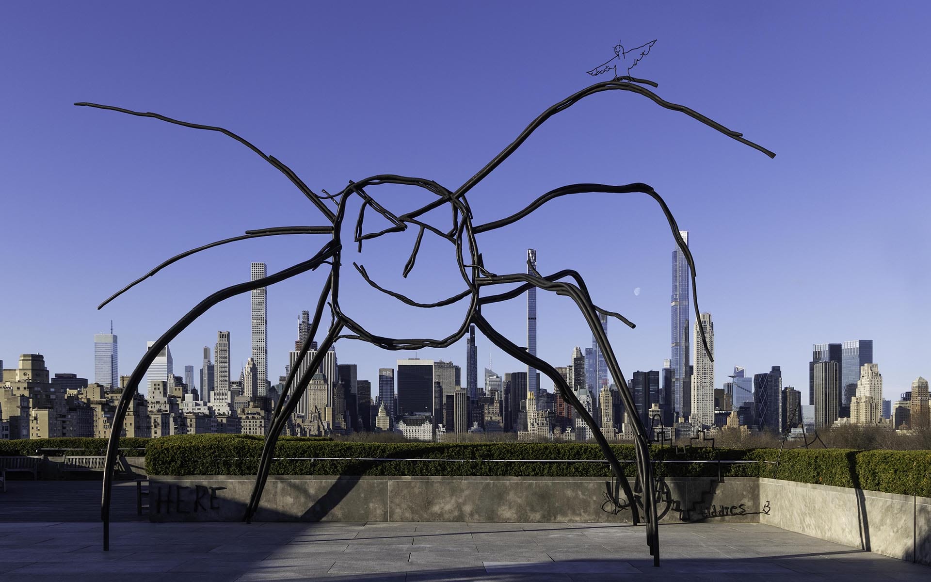 Spider-shaped sculpture against a city skyline background, with tall buildings under a clear blue sky.