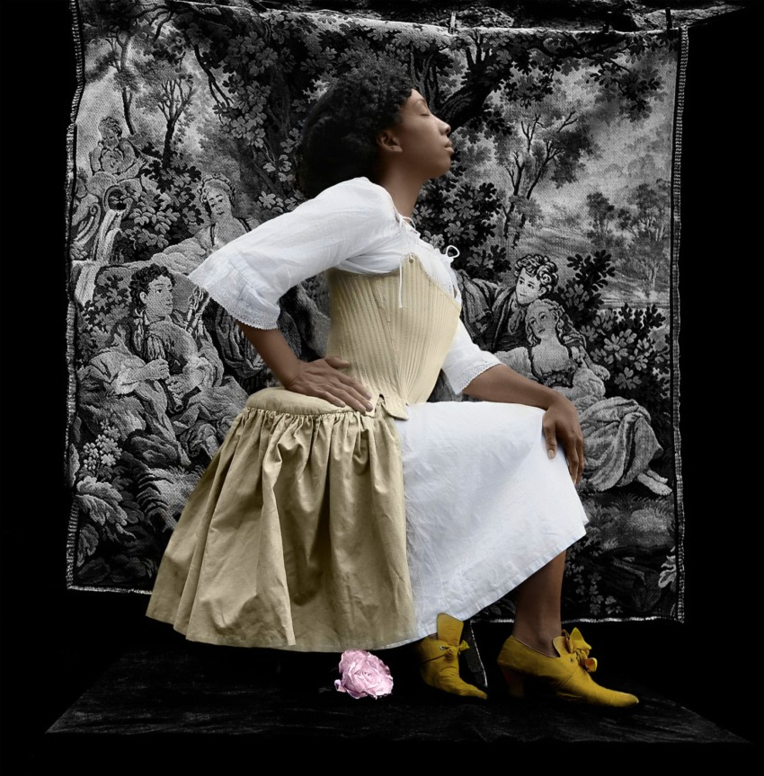 Person in historical costume posing against a vintage tapestry backdrop, wearing a corset, skirt, and yellow shoes.