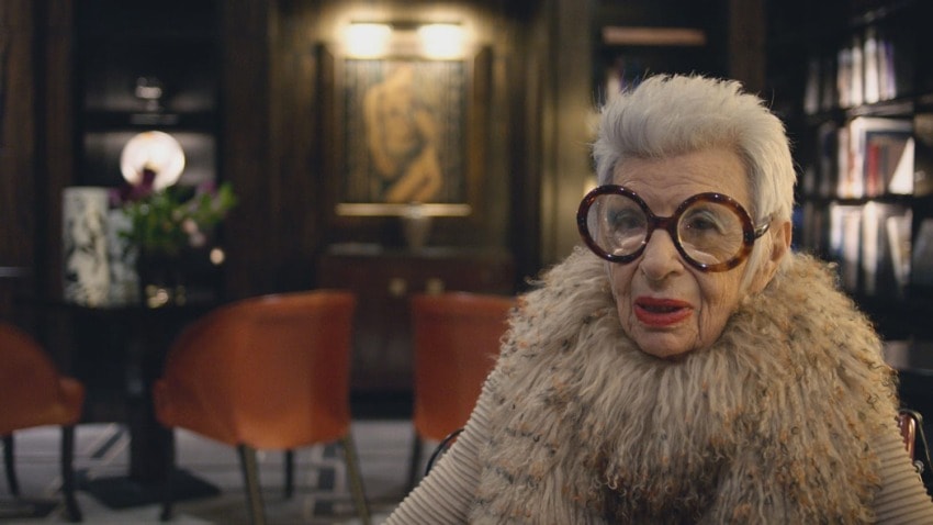 Elderly woman with grey hair and large glasses, wearing a fur collar, sitting in a dimly lit room with red chairs and artwork.
