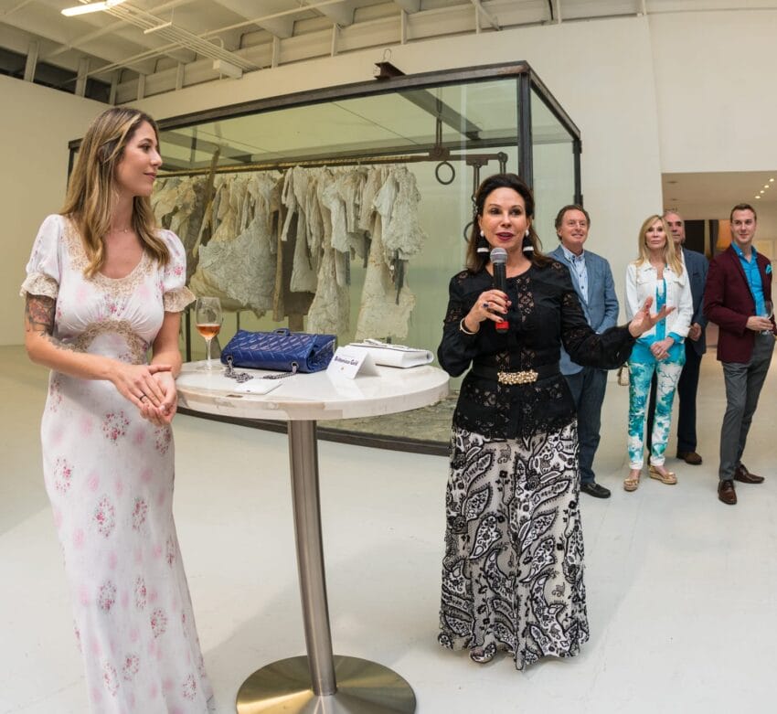 Woman speaks at an art gallery event with a small audience, featuring a textile and phone display on a table.