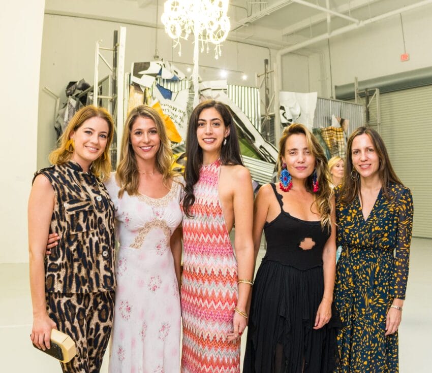 A group of five women posing together in a brightly lit modern art gallery setting.