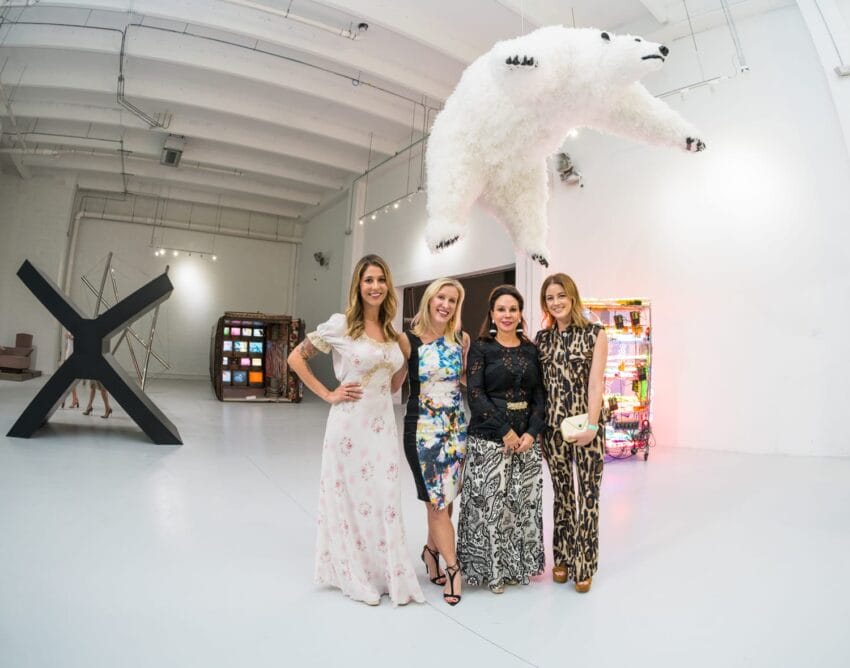 Four women posing in an art gallery with a polar bear sculpture hanging from the ceiling and various art pieces around.