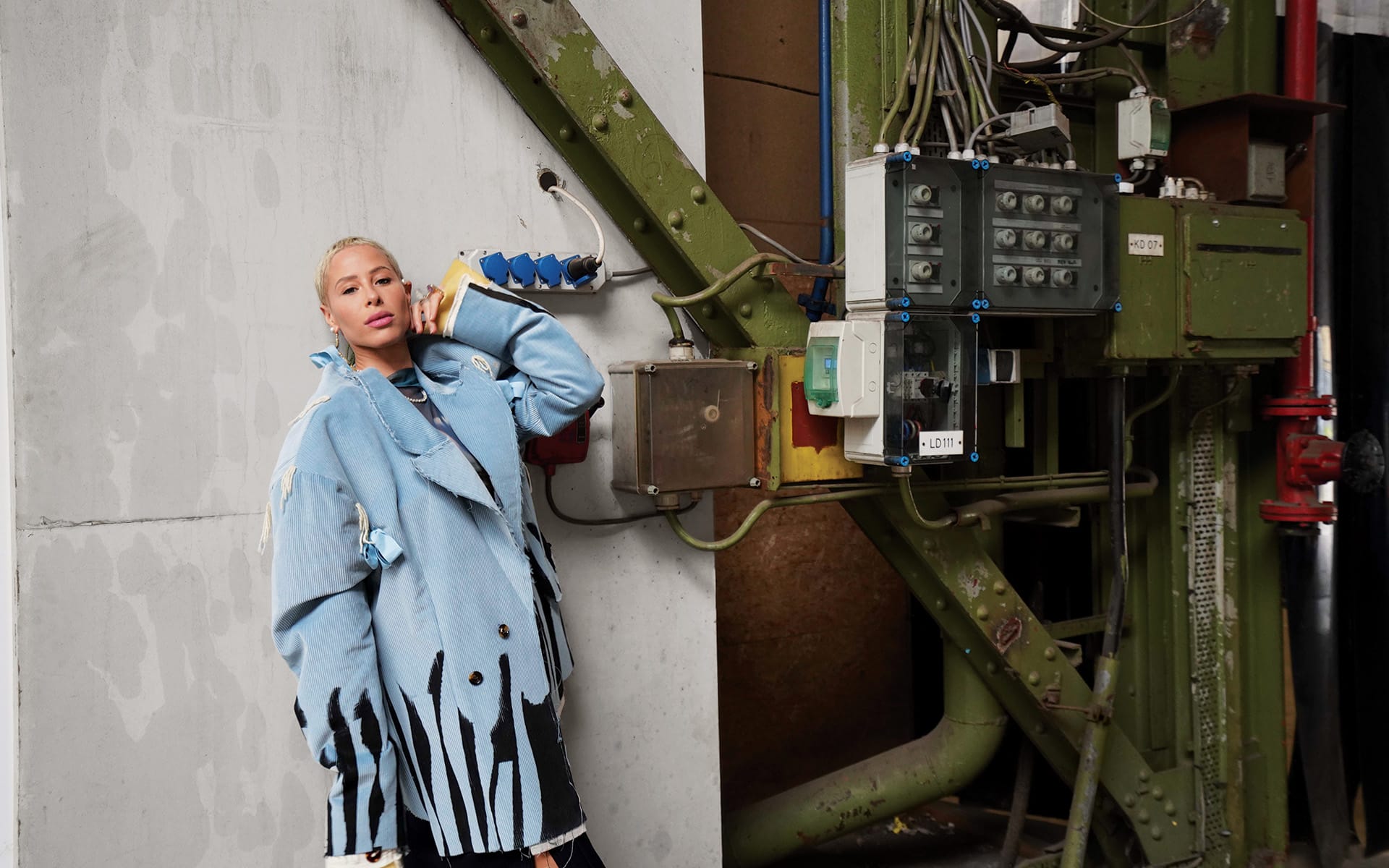 Person in a blue coat with a phone, standing next to industrial equipment with wires and panels.