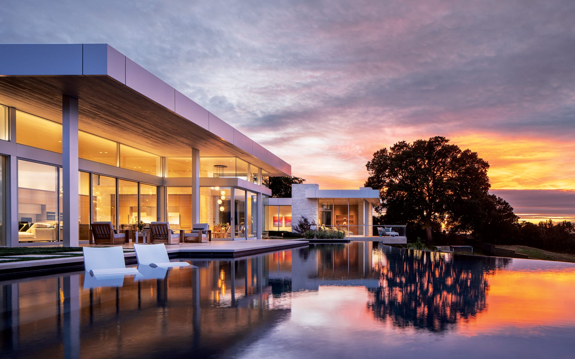 Modern luxury house with large glass windows, pool reflecting a vibrant sunset sky, and trees in the background.