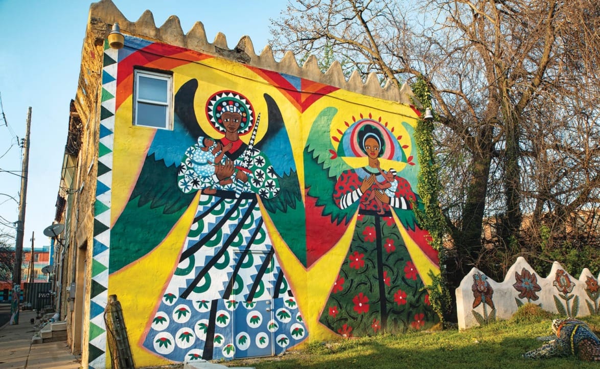 Colorful mural of two angels with vibrant wings and intricate patterns on a building wall, surrounded by trees.