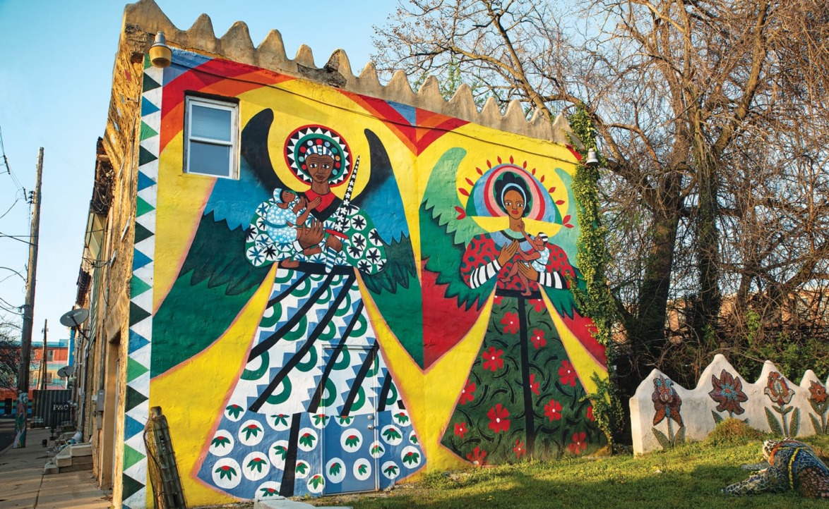 Colorful mural of two angels with vibrant wings and intricate patterns on a building wall, surrounded by trees.