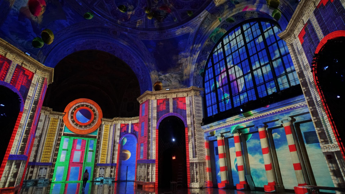 Colorful digital art projected on interior walls and ceiling of a grand architectural hall with columns and arches.