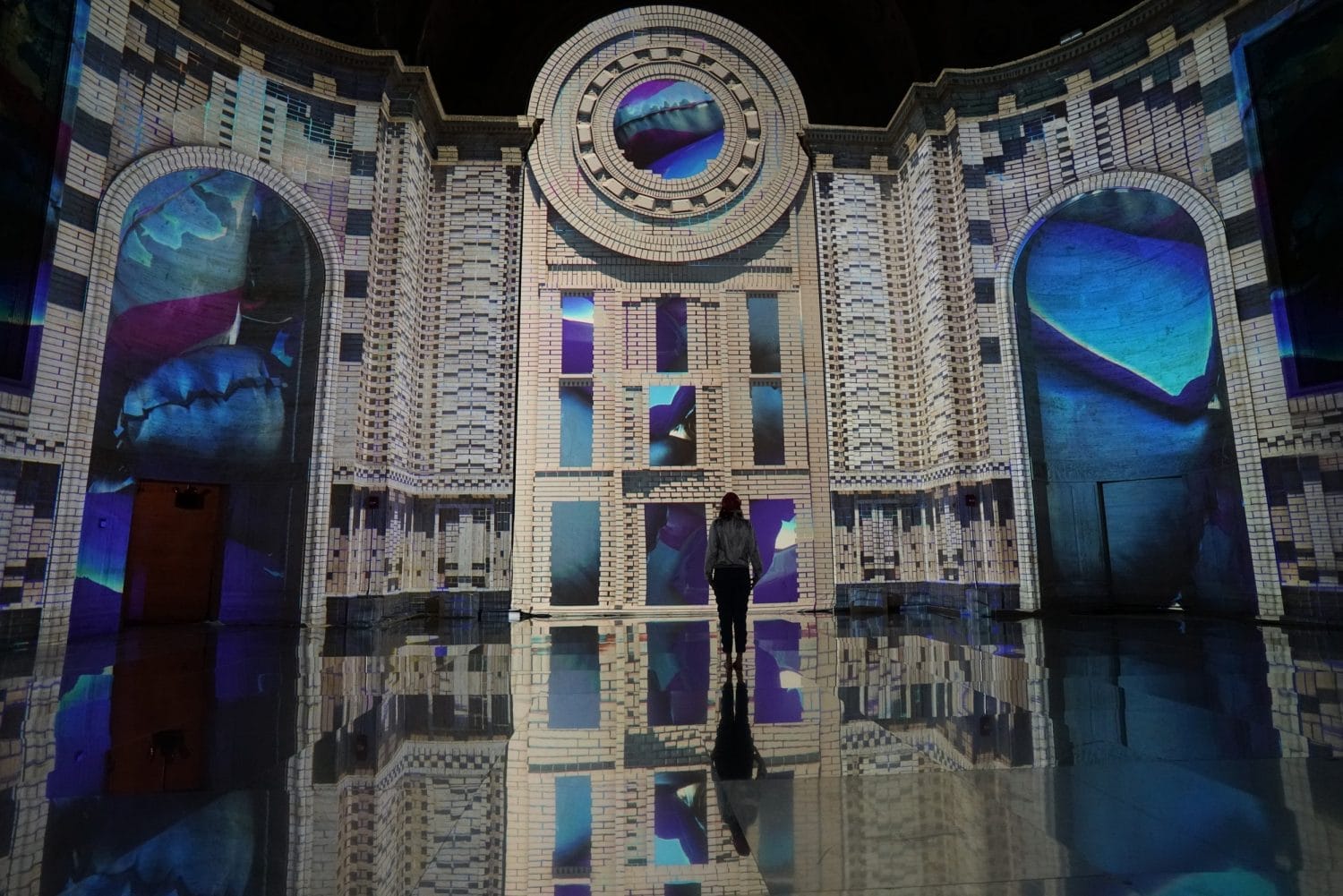 Person standing in a large room with intricate digital projections on walls and reflective floors.