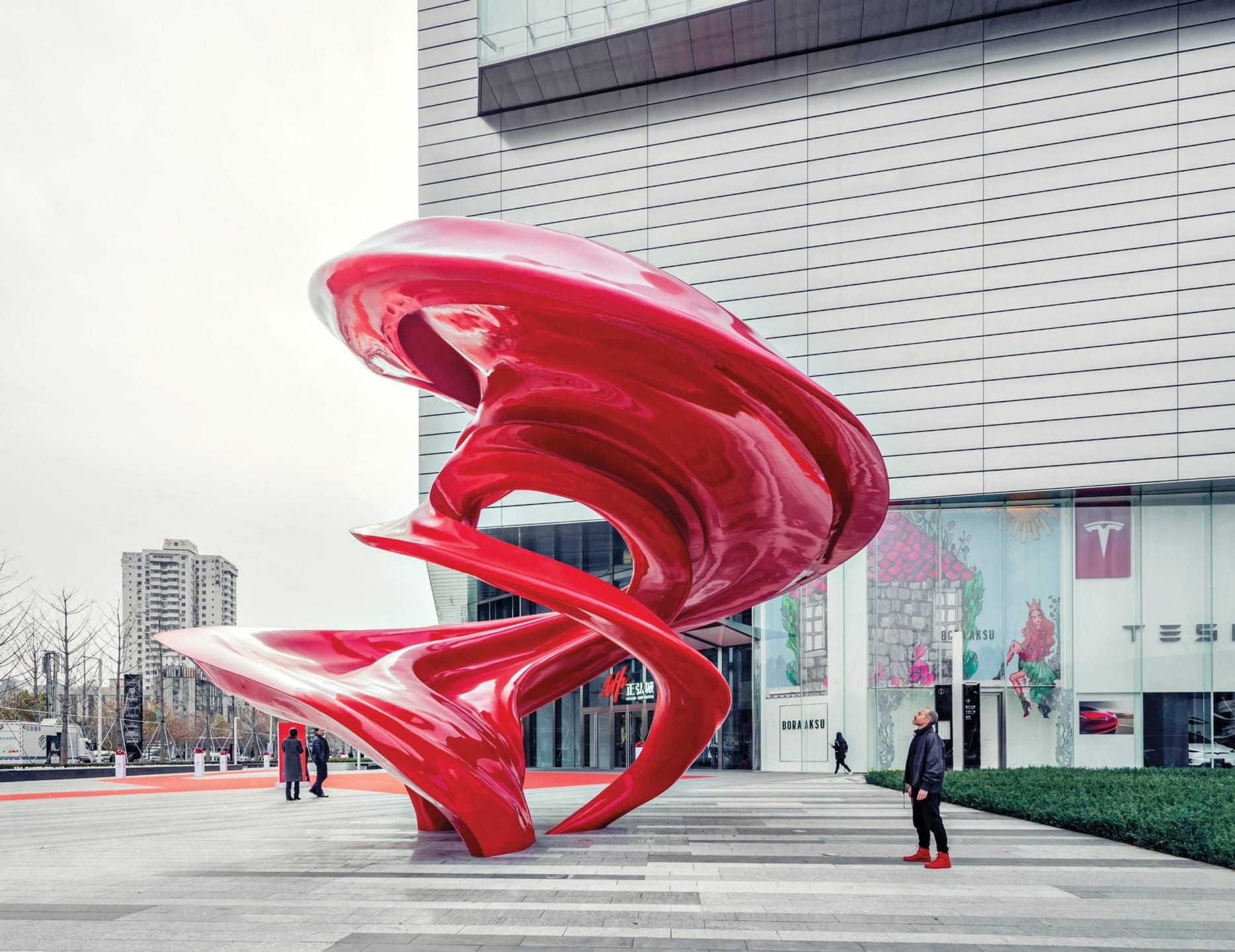 Large red abstract sculpture in urban plaza with modern building backdrop and people nearby.