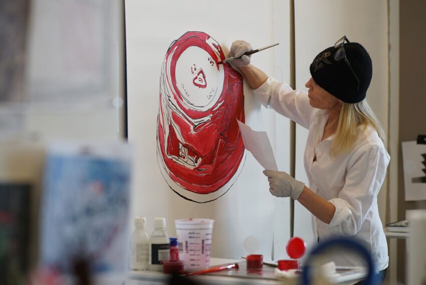 Artist painting a large red and white stylized aluminum can on a canvas, wearing a black beanie and white shirt.
