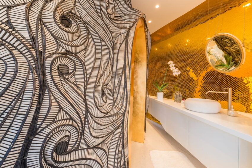 Luxurious bathroom with gold mosaic wall, black and white patterned partition, white vanity, and circular mirror.