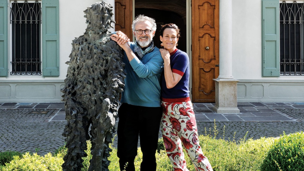 Two people smiling and posing beside a textured statue in front of a building with wooden doors and green shutters.