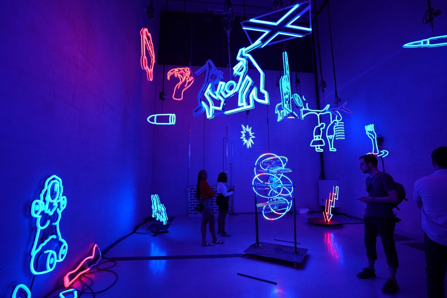 People viewing colorful neon light art installation in a dimly lit room with various abstract shapes and figures.