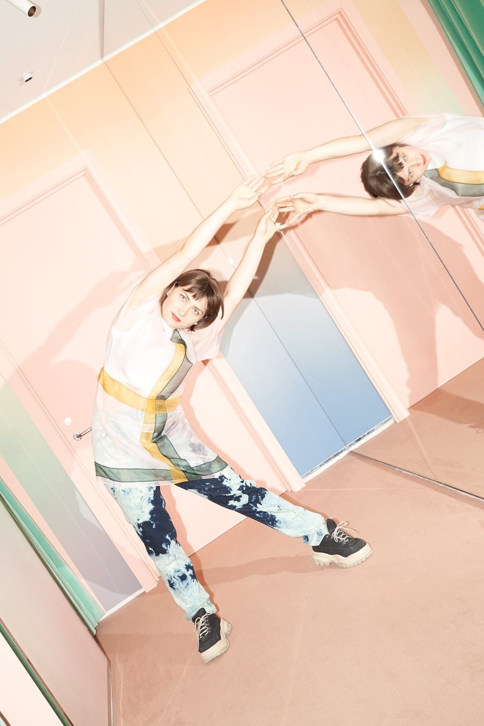 Person stretching in front of mirrored wall, wearing a patterned outfit with tie-dye pants and sneakers in a colorful room.