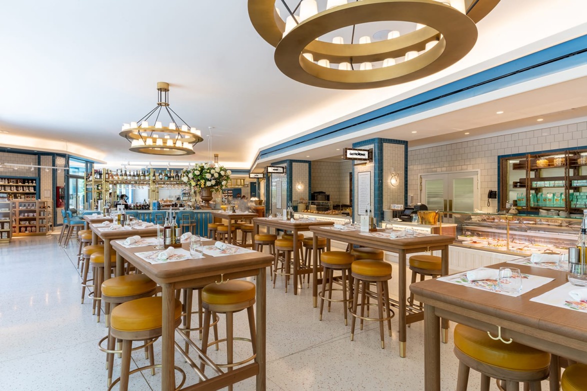 Spacious modern restaurant interior with high tables, wooden chairs, chandeliers, and a well-stocked bar in the background.