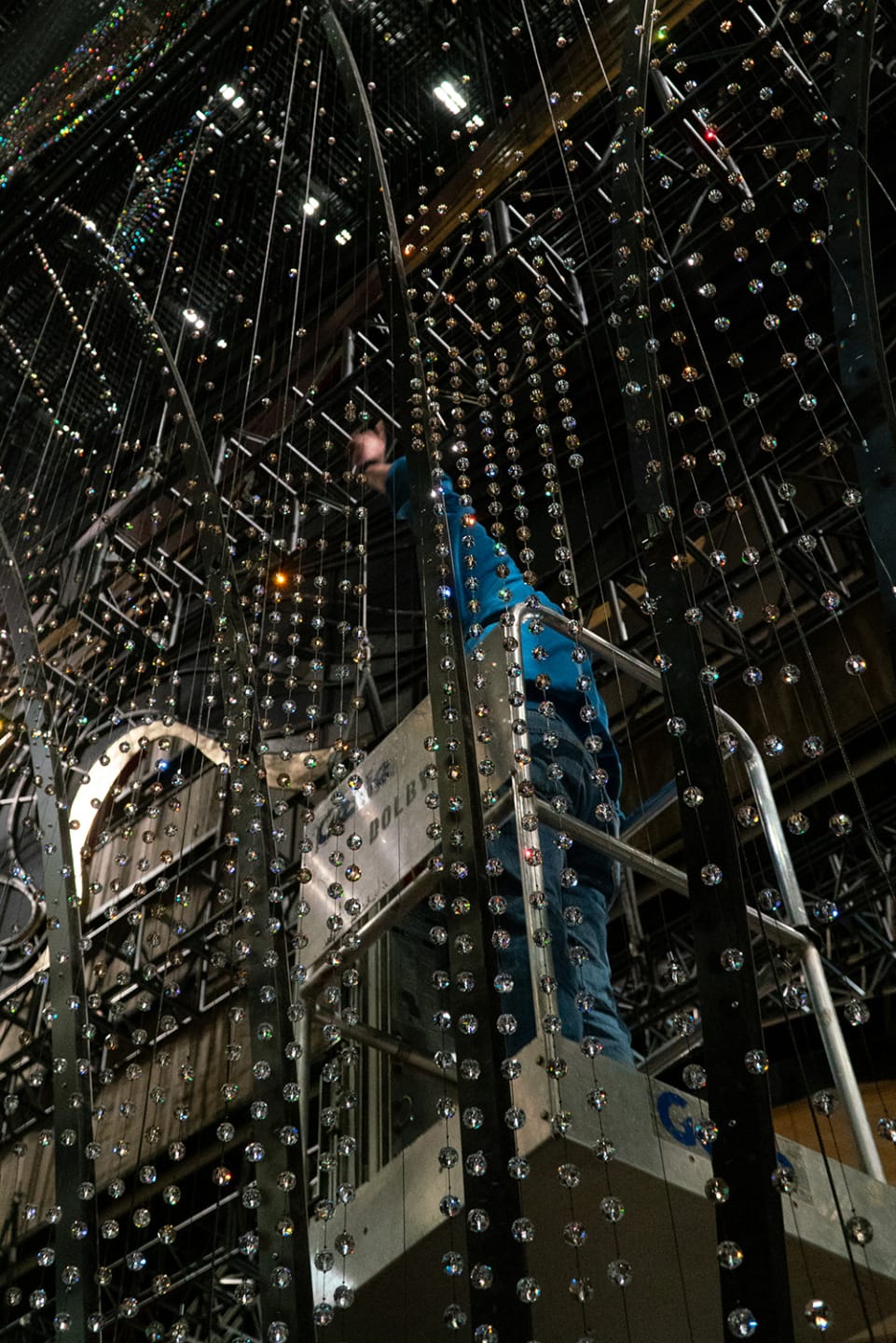 Person standing on a lift behind a curtain of strings with beads, surrounded by industrial metal framework.