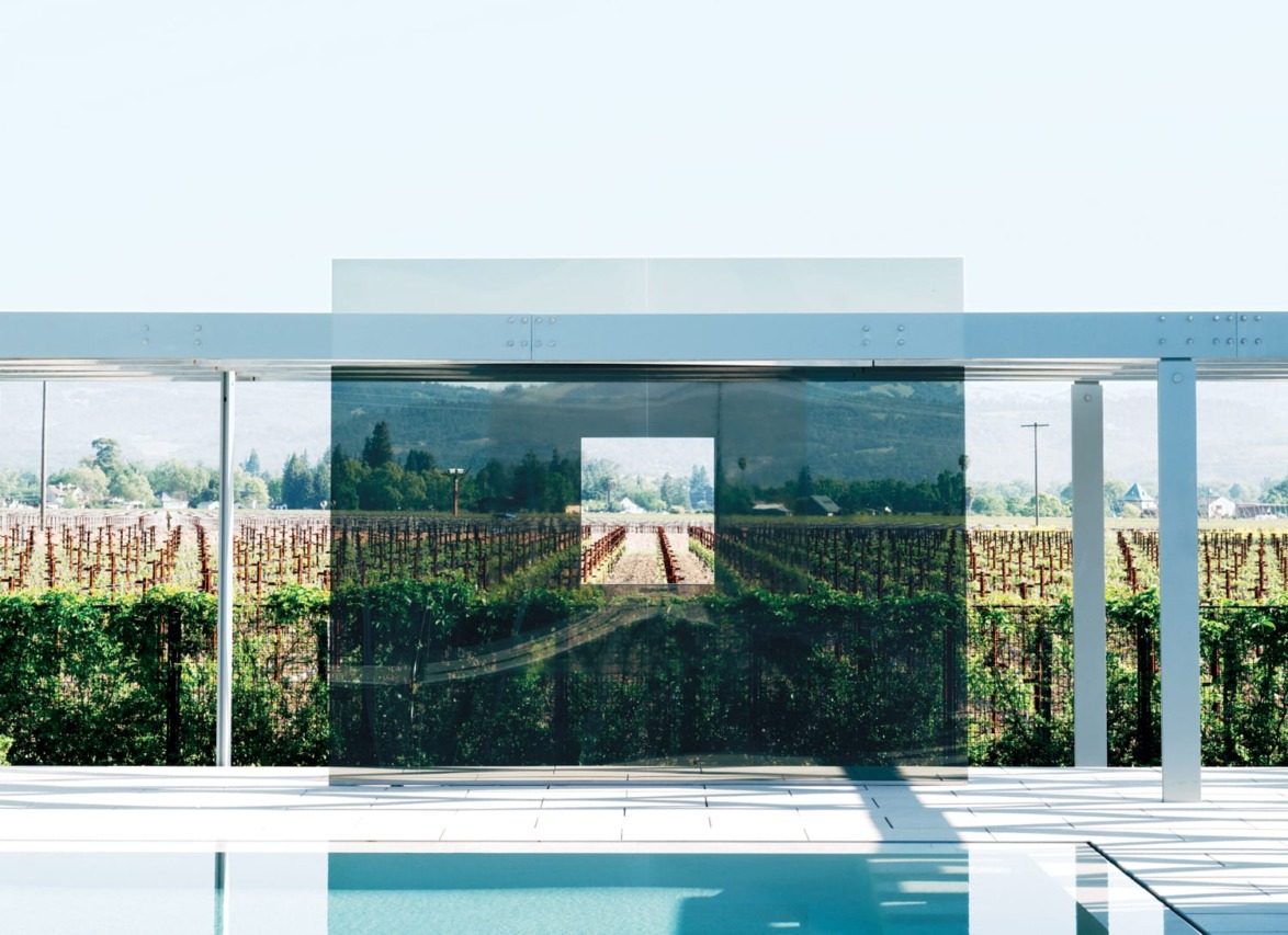 Modern art installation with glass panels reflecting vineyard landscape, framed by a sleek metal structure and pool.