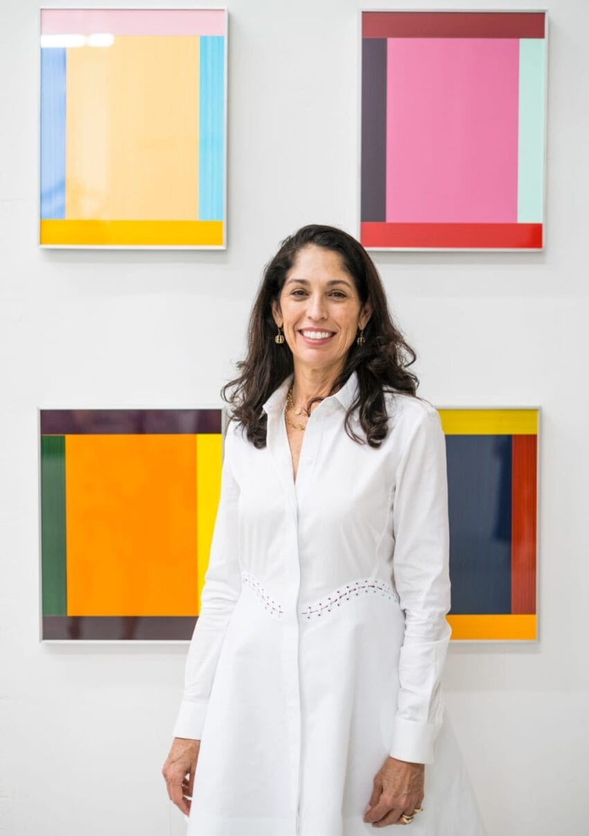 Person in white dress standing in front of colorful abstract paintings on a white wall.