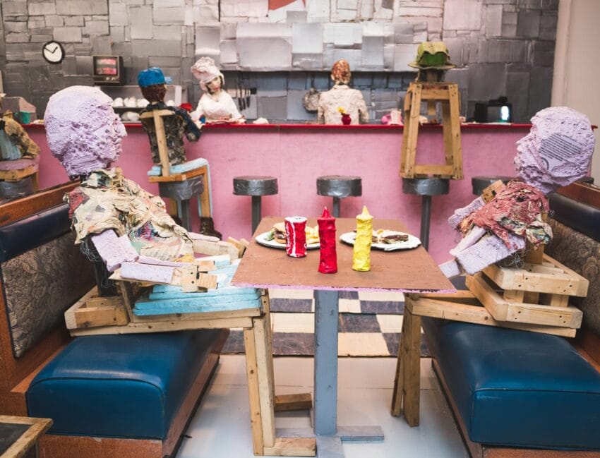 Art installation of figures made from recycled materials sitting at a diner with a pink counter and condiments on the table.