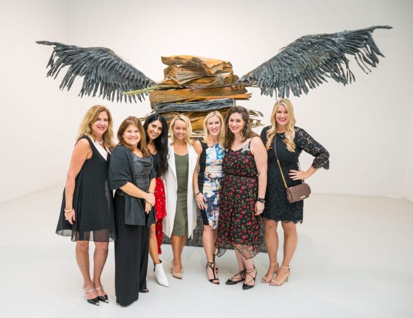 Seven women posing in front of a large metal sculpture with wings in an art gallery setting.