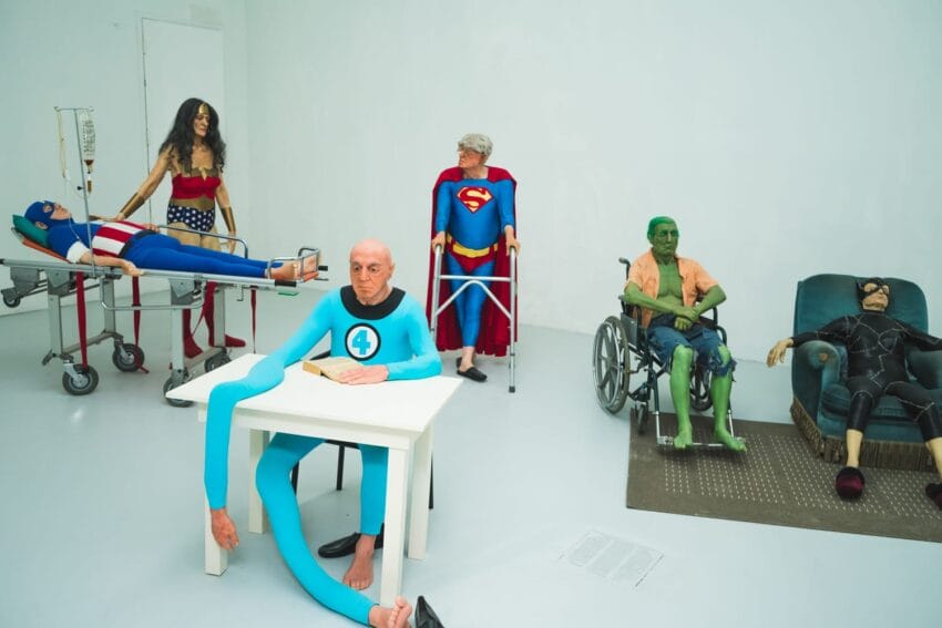 Retired superheroes in costumes, including a stretchered Captain America and a seated Hulk, gather in a white room.