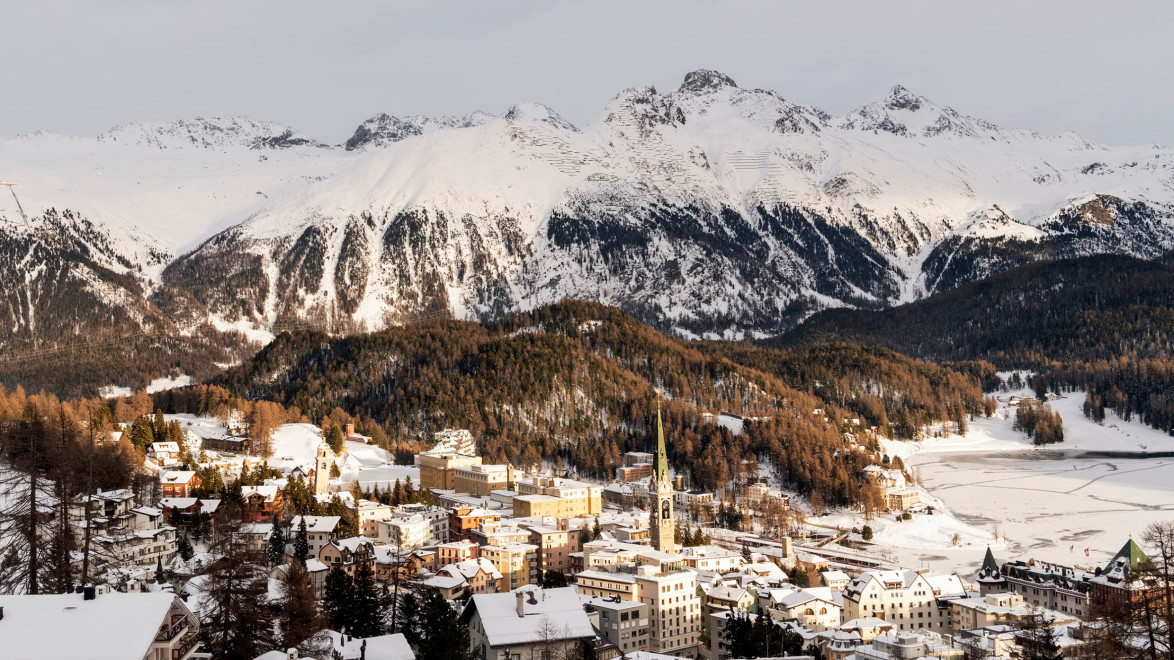 Snow-covered mountain village with trees and church nestled in a valley, surrounded by majestic peaks under a cloudy sky.