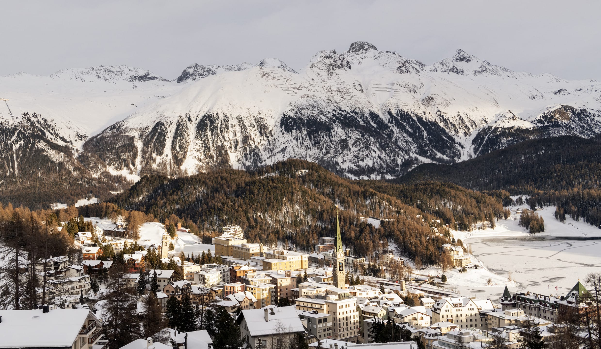Snow-covered mountain village with trees and church nestled in a valley, surrounded by majestic peaks under a cloudy sky.