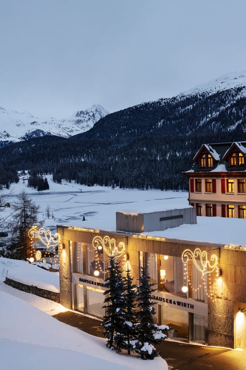 Winter scene with snow-covered mountains, lit buildings, and pine trees in front of an art gallery, under a cloudy sky.